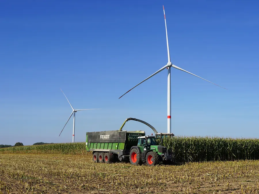 Häcksler und Schlepper mit Ladewagen am Maisschlag, dahinter zwei Windräder